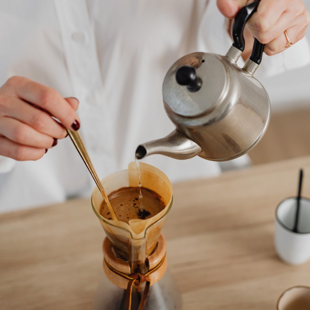 Person pouring coffee from a silver teapot into a filter basket on a wooden surface.