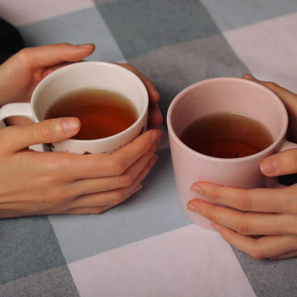 Two hands holding mugs of tea on a checkered tablecloth